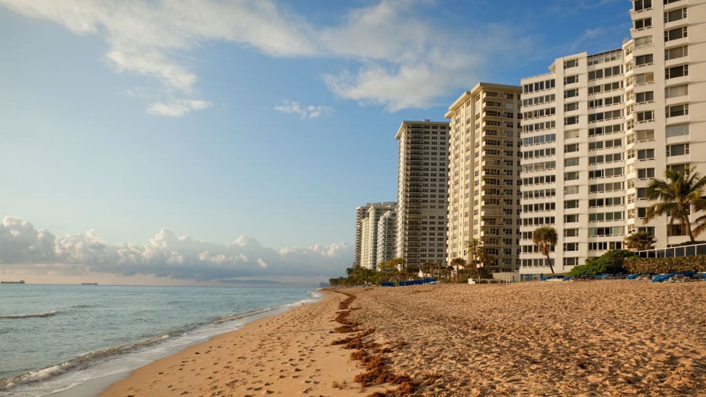 High-rise beachfront condominiums line a sandy beach with gentle waves and scattered seaweed under a partly cloudy sky.